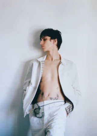 A stylish young adult stands in a white jacket, light pants, and a pendant necklace against a pale background, capturing a fashion-forward moment.