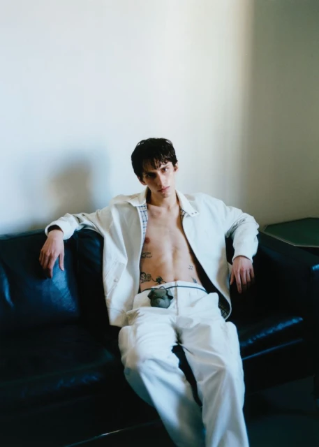 Adult male fashion model with exposed chest and tattoos sitting on a black leather couch in front of a white wall in a studio.