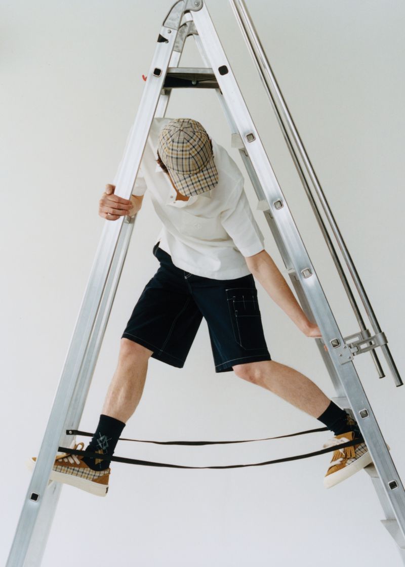 Young adult climbs a silver aluminum ladder indoors. He wears dark shorts, a white t-shirt, a patterned cap, and tan shoes.