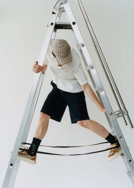 Young adult climbs a silver aluminum ladder indoors. He wears dark shorts, a white t-shirt, a patterned cap, and tan shoes.