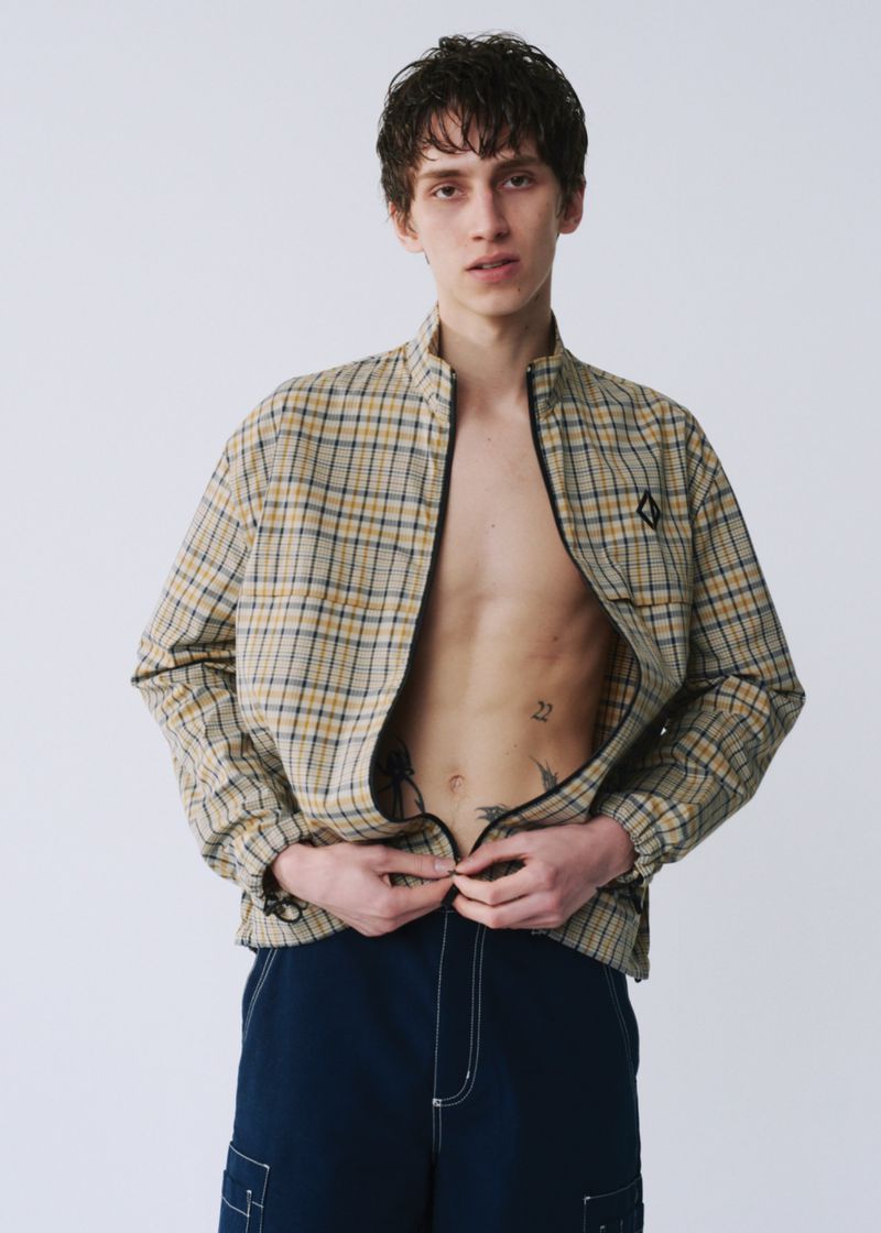 Young man in open plaid jacket and blue pants, unzipping jacket in studio. Neutral backdrop, casual fashion for editorial or lifestyle.