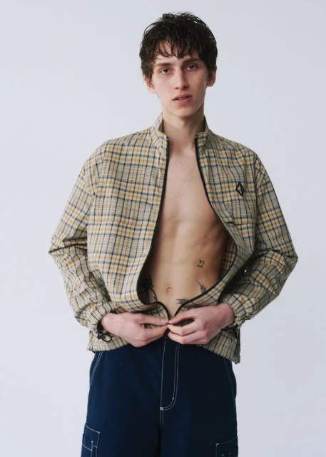 Young man in open plaid jacket and blue pants, unzipping jacket in studio. Neutral backdrop, casual fashion for editorial or lifestyle.