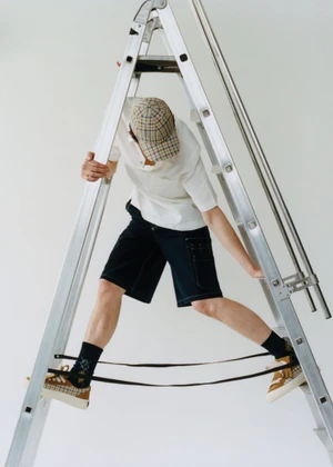 Man wearing a plaid cap, white polo shirt, and black shorts posing amidst a large metal ladder against a plain white background.