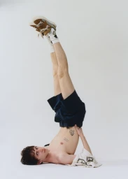 A man lying upside down with legs raised straight up wearing dark shorts, white socks, and brown sneakers against a white background.