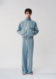 A male model wearing a light blue tracksuit set standing against a plain white studio backdrop.