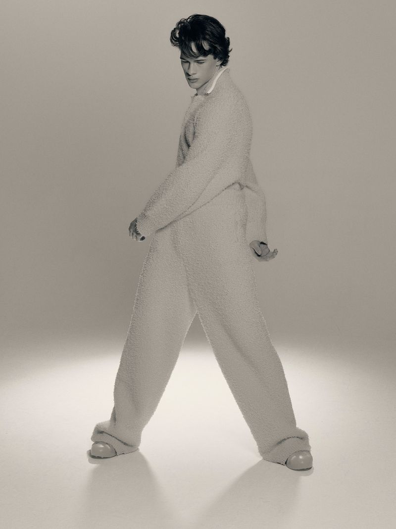 Young adult male fashion model poses in a soft, cream-colored, plush-textured lounge outfit, angled in a studio setting.