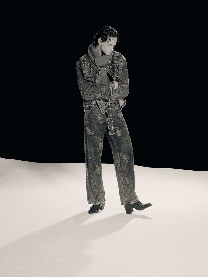 Monochrome full shot of a young adult man in a patterned denim jacket and jeans, posing in a studio with a black and white backdrop.