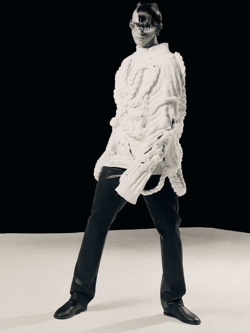 Monochromatic fashion shot of a slender adult posing in an extravagant knit sweater, black pants, and fashion eyewear, against a stark black backdrop.