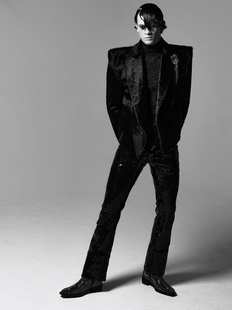 Full-length studio shot of a male model wearing a textured black suit, textured turtleneck, and black boots on a neutral background.