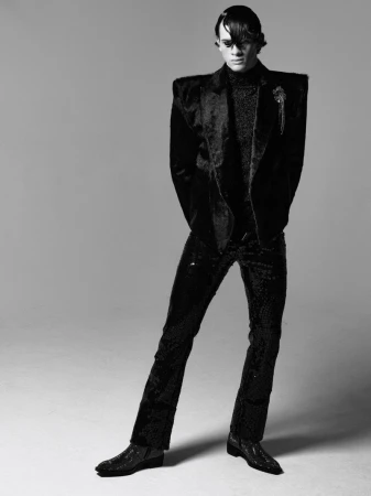Full-length studio shot of a male model wearing a textured black suit, textured turtleneck, and black boots on a neutral background.