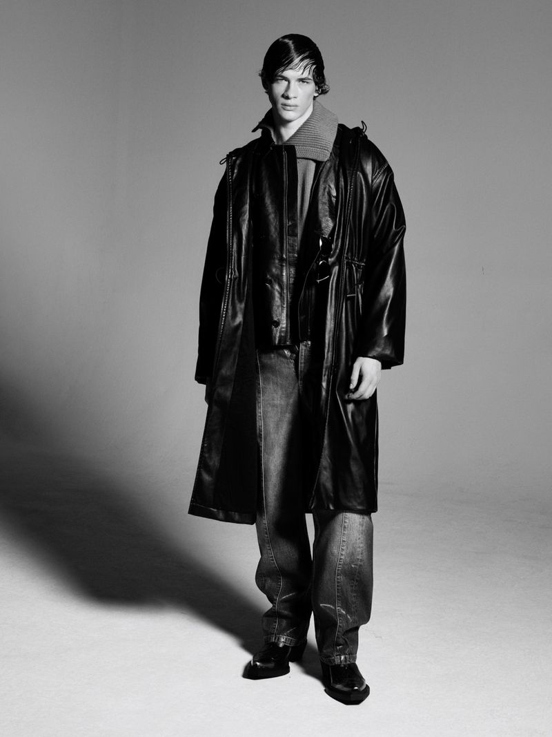 Black and white portrait of an adult male fashion model wearing a black overcoat, grey hooded sweatshirt, and blue jeans in a photo studio.