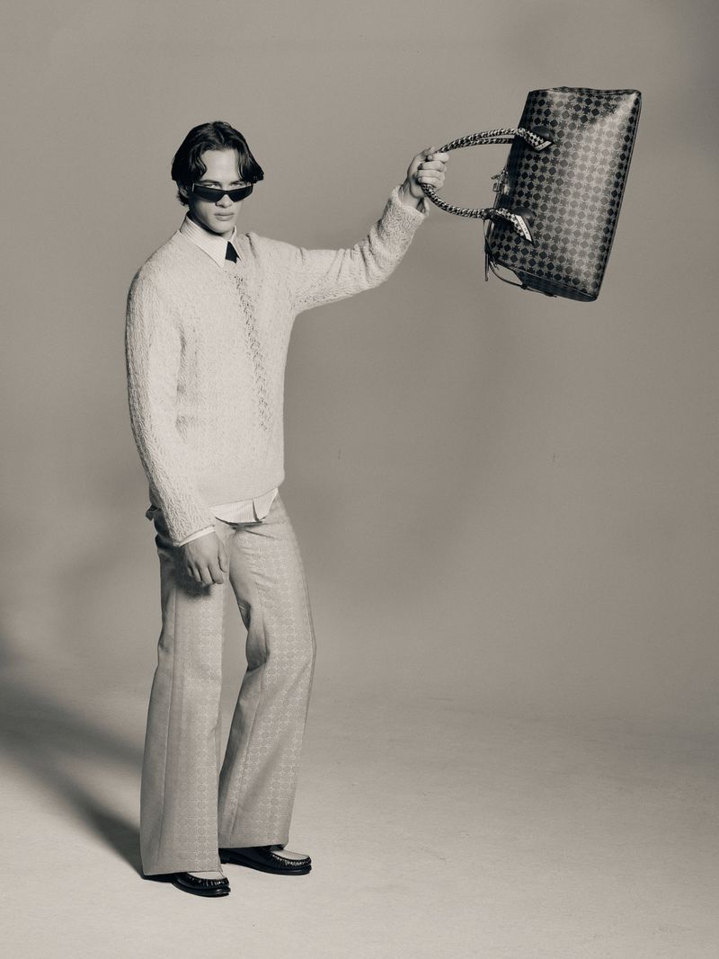 Monochrome full shot of a man in a sweater and wide-leg pants holding a checkered tote bag against a neutral background in a studio.