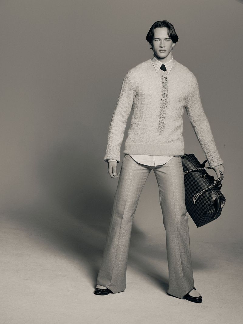 Studio shot of a slender man in a cream textured sweater, light patterned pants, and carrying a black duffel bag.