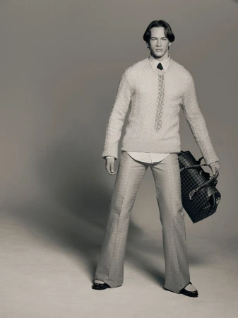 Studio shot of a slender man in a cream textured sweater, light patterned pants, and carrying a black duffel bag.