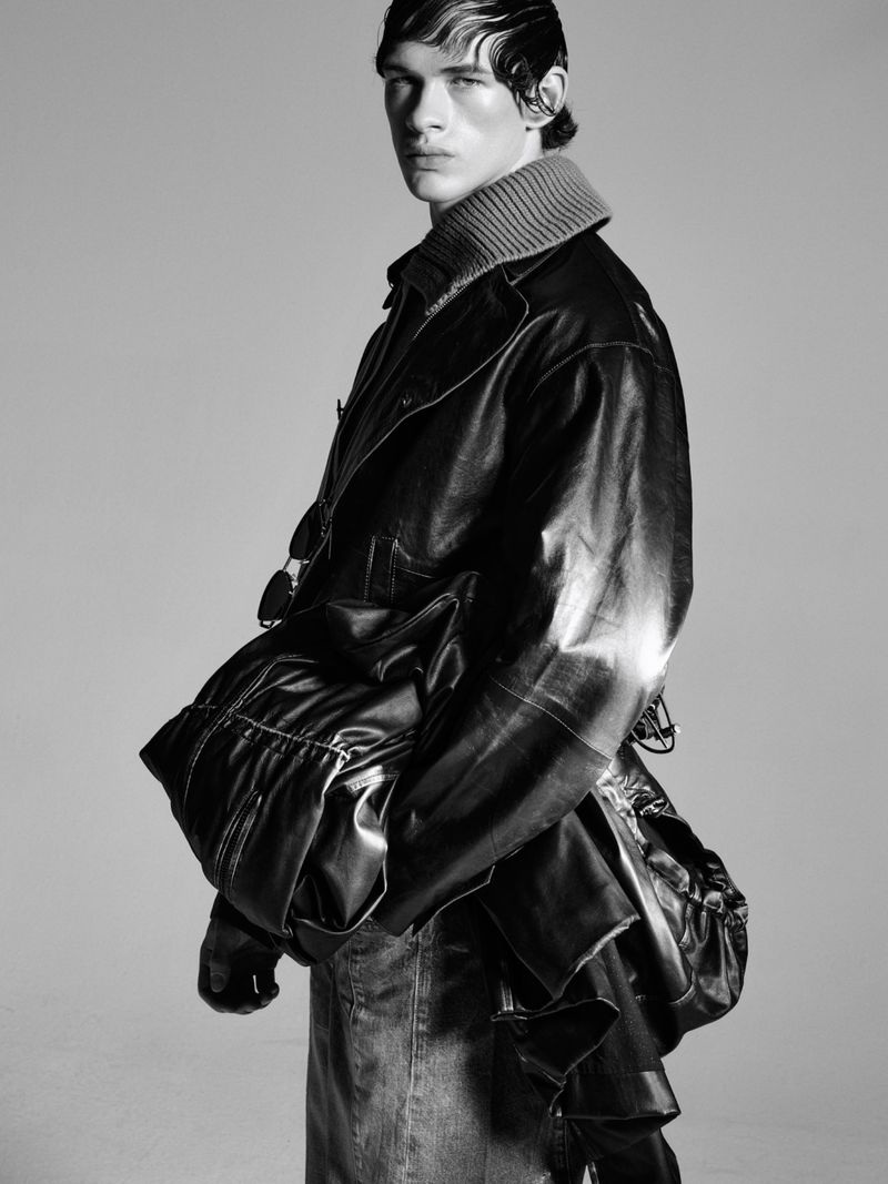 Monochromatic fashion shot of a fair-skinned male model wearing a dark jacket and denim skirt in a studio with a neutral background.