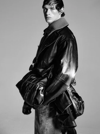 Monochromatic fashion shot of a fair-skinned male model wearing a dark jacket and denim skirt in a studio with a neutral background.