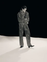 A young male model wearing patterned denim jacket and trousers posing against a dark and light studio background.