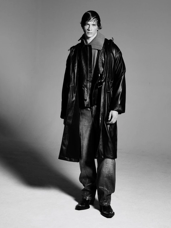 A young male model wearing a black leather trench coat, layered collar, and dark denim jeans against a seamless grey backdrop.