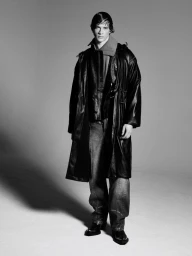 A young male model wearing a black leather trench coat, layered collar, and dark denim jeans against a seamless grey backdrop.