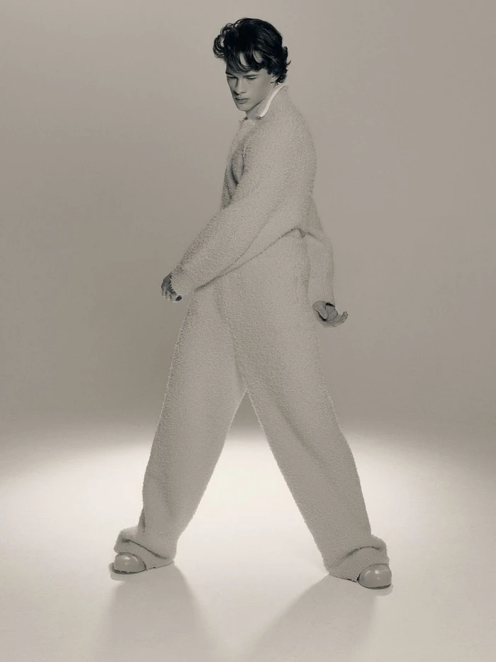 A man wearing a fuzzy matching sweater and trousers standing against a seamless pale studio background.