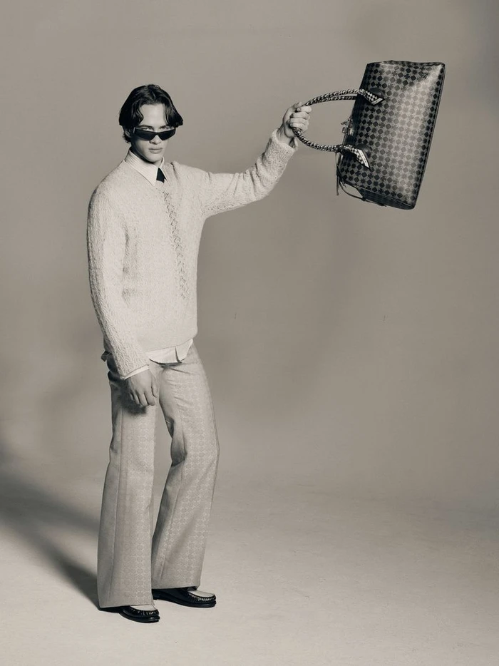 Man wearing sunglasses and a textured sweater holding up a patterned handbag in a studio