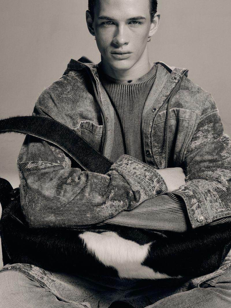 A young male model with crossed arms wearing a textured jacket against a plain grey background