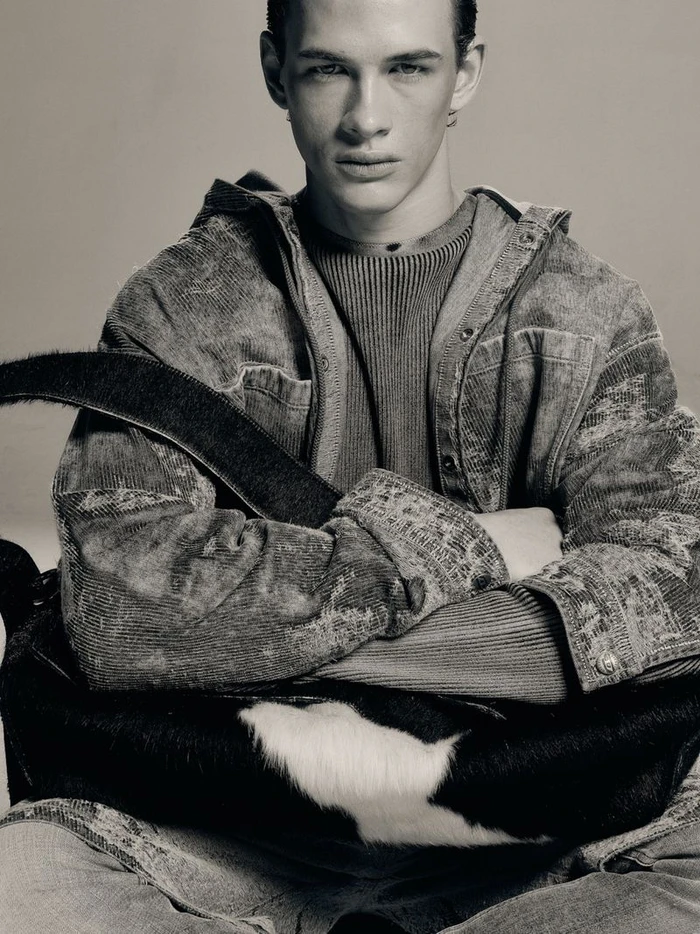A young male model with crossed arms wearing a textured jacket against a plain grey background