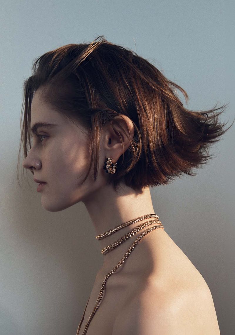 Profile portrait of a young adult female with short brunette hair wearing gold jewelry against a neutral background.