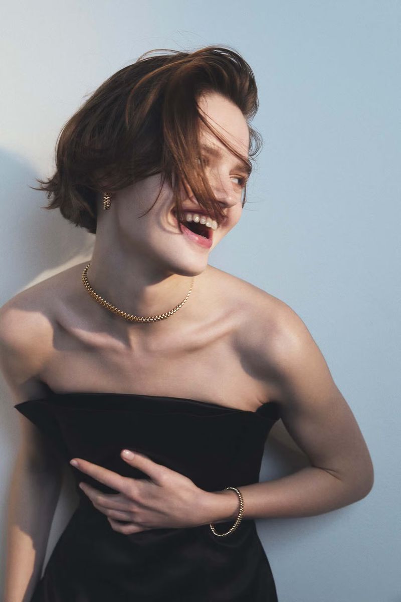 A smiling woman with short brown hair wearing a black strapless dress and gold necklace and bracelet posing in front of a wall.
