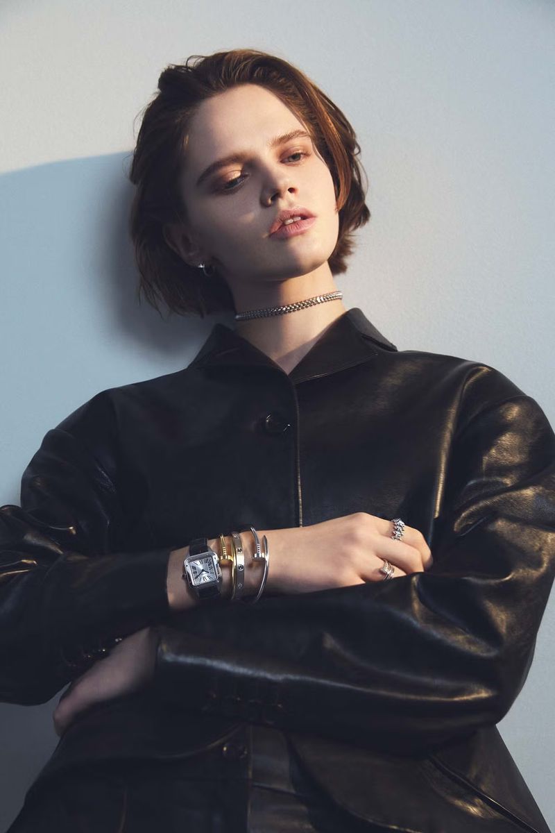 Portrait of a woman in a black leather jacket with silver jewelry, standing against a soft blue wall with natural window lighting.