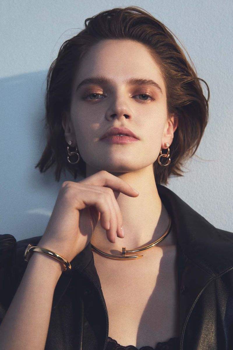 Close-up of a young woman with short hair wearing jewelry, including a necklace, earrings, and a bracelet, with a light blue background.