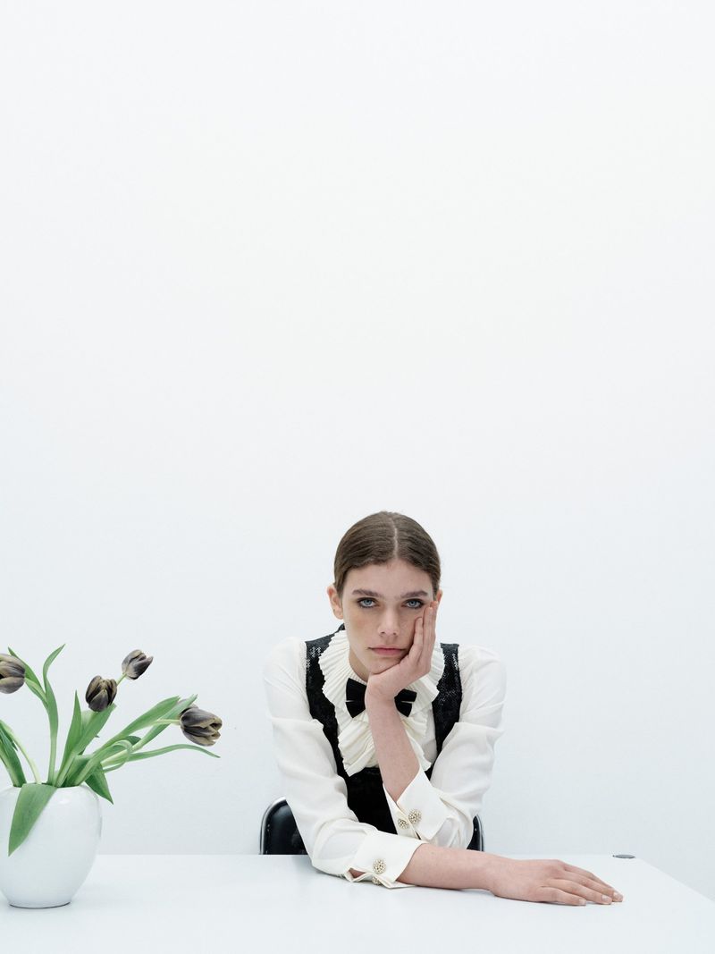 A young woman with dark hair and fair skin poses at a table with dark tulips in a white vase against a minimalist white backdrop.
