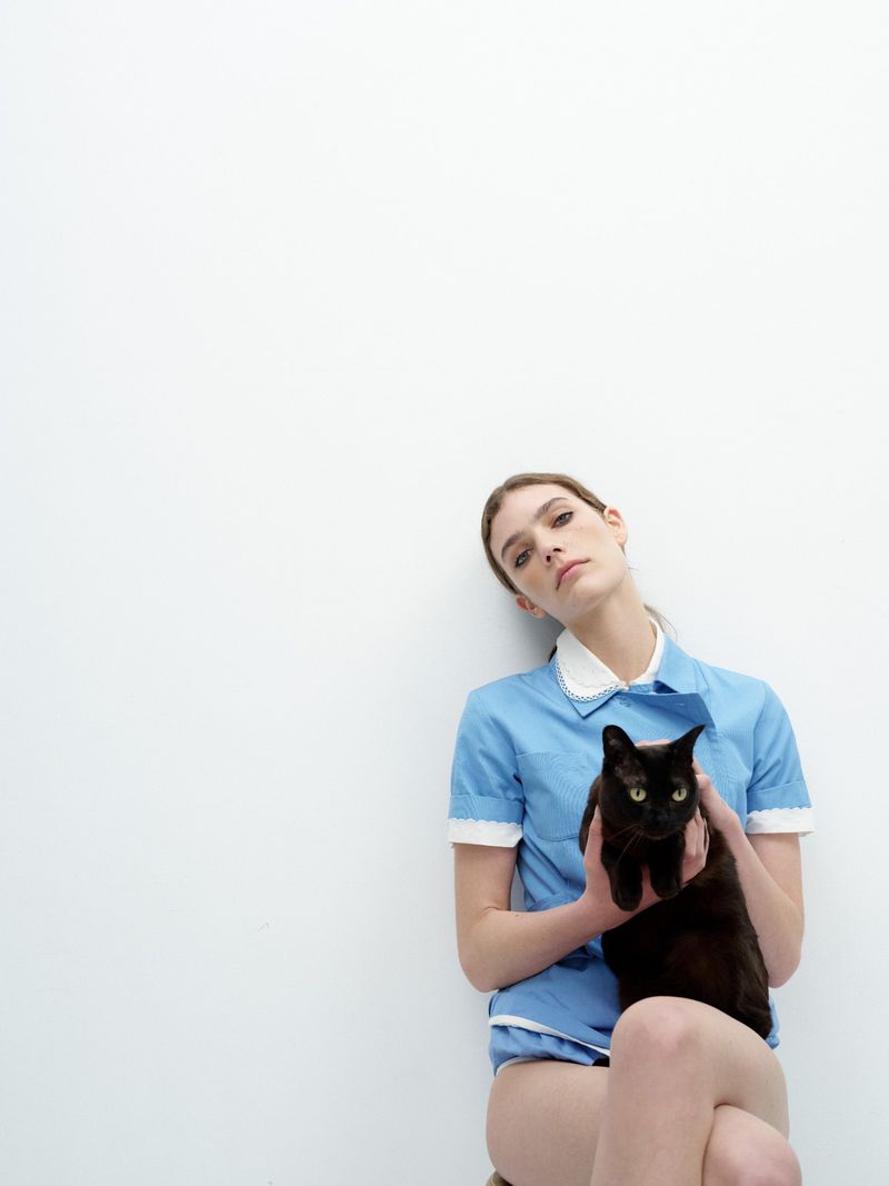 Woman in blue uniform poses with a black cat against a white background in a studio. The portrait is shot at eye-level.
