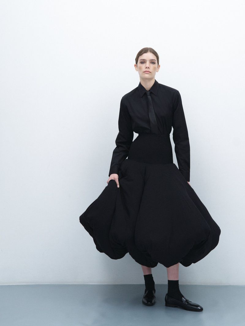 A fashionable woman in a black shirt, tie, and full skirt poses in a studio, holding the skirt, against a white wall, full shot, eye-level.