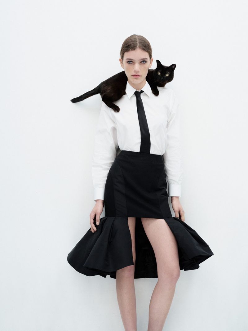 Fashion shot of a woman in a white shirt and black skirt, posing with a black cat draped over her shoulders in a bright photo studio.
