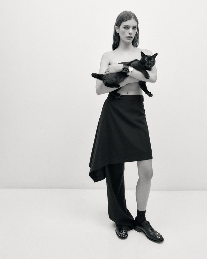 A person in a dark skirt and trousers holds a black cat in a studio setting, photographed in black and white with even lighting.
