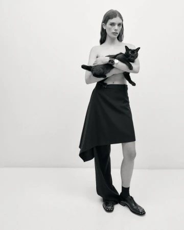 A person in a dark skirt and trousers holds a black cat in a studio setting, photographed in black and white with even lighting.