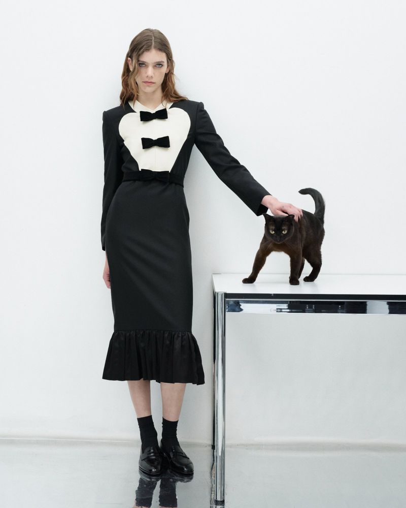 Fashionable woman in black dress with black cat standing on a table in a studio. The background is white and reflective.