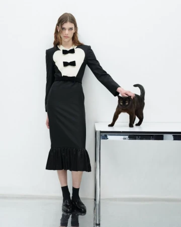 Fashionable woman in black dress with black cat standing on a table in a studio. The background is white and reflective.