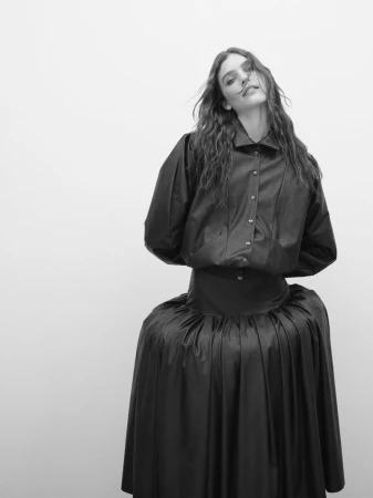 Model Betsy Gaghan wearing a black buttoned shirt and flared skirt against a solid white background.