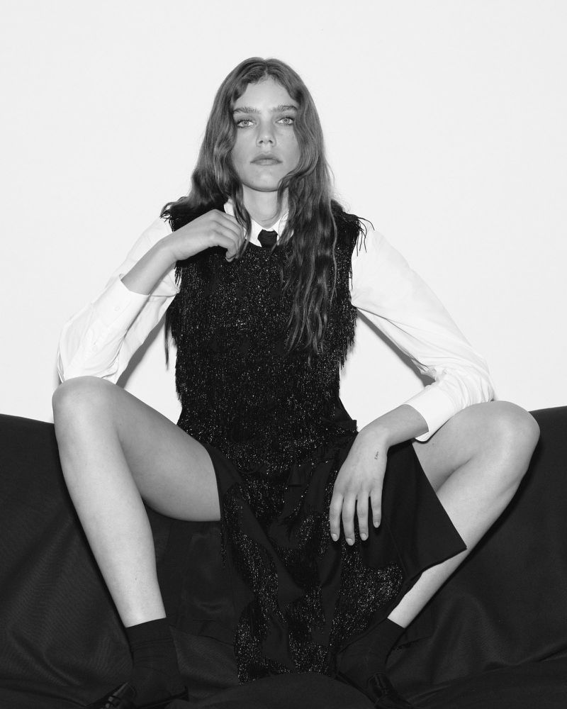 A woman sitting on a black couch wearing a white dress shirt and a black sparkle vest, with long brown hair and black shoes.