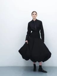 Model Betsy Gaghan in a structured black shirt and voluminous skirt against a seamless white studio background.