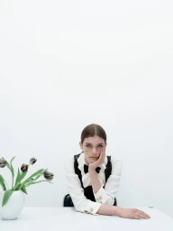 Model Betsy Gaghan seated at a white table with a vase of tulips against a plain white seamless studio background.