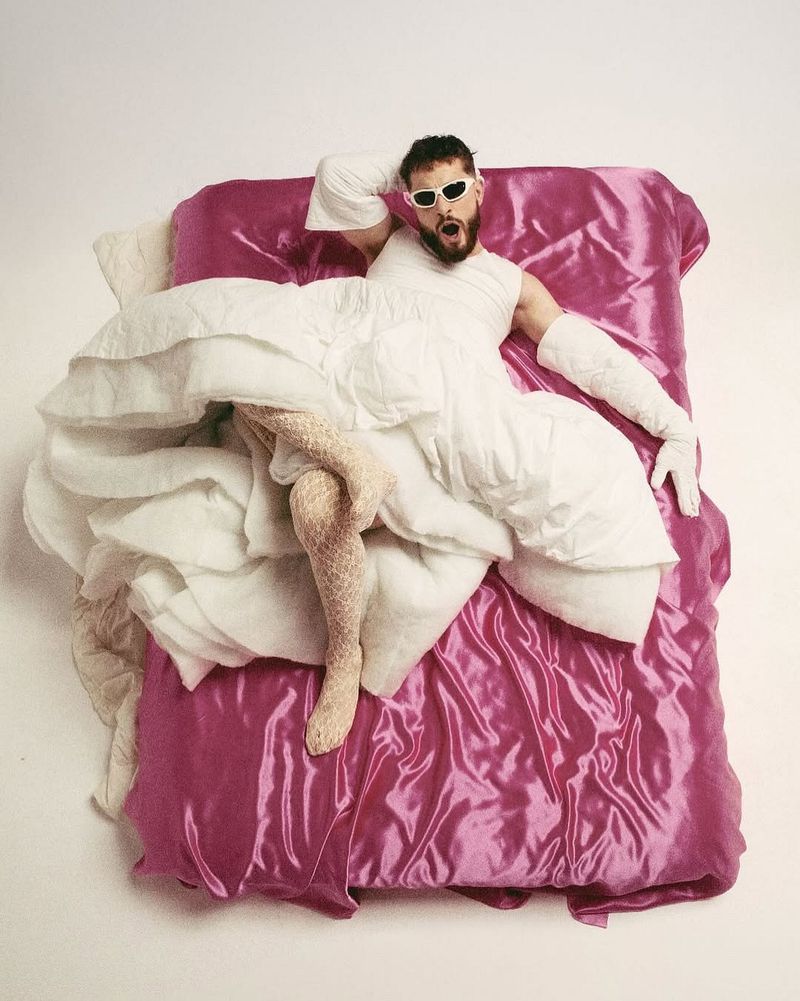 A man lies on a pink satin bed covered in a white quilted blanket, wearing a white shirt, fishnet stockings, and white sunglasses.