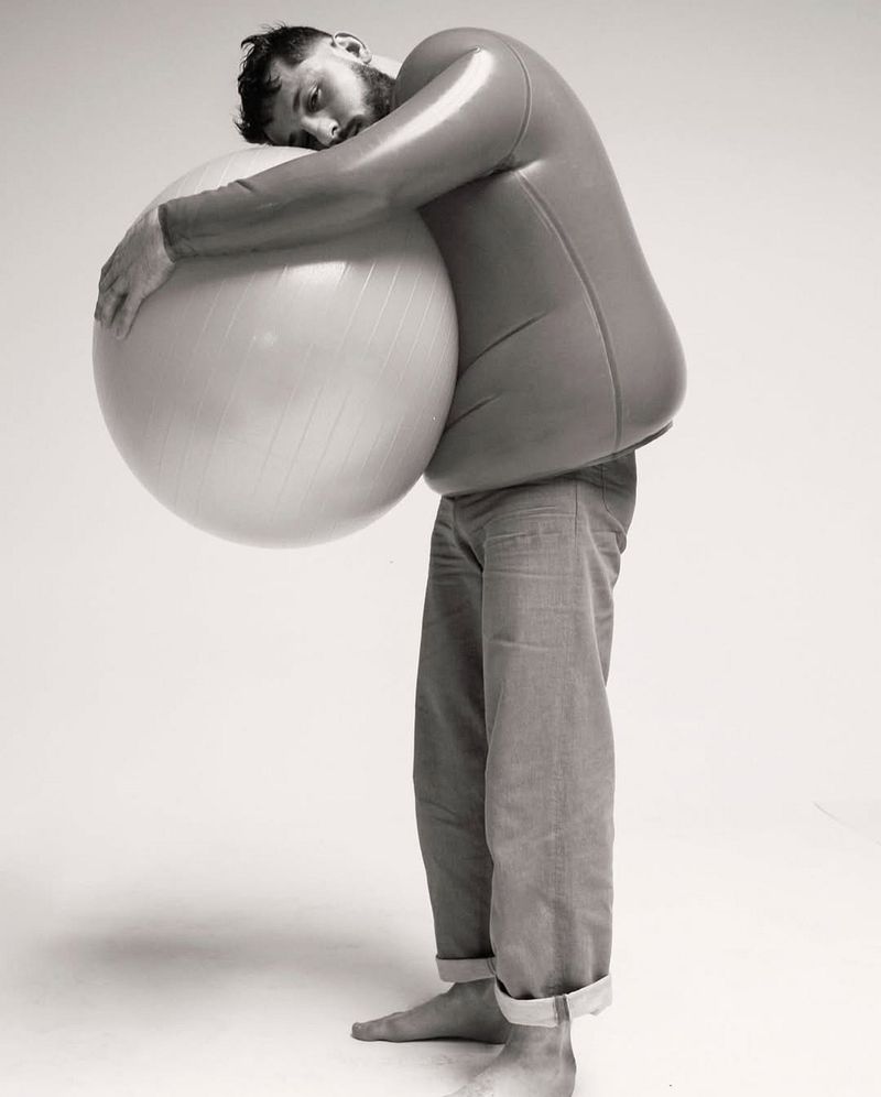 Adult male embracing exercise ball in studio setting. He is wearing a skin-tight top, jeans, and is barefoot against a plain background.