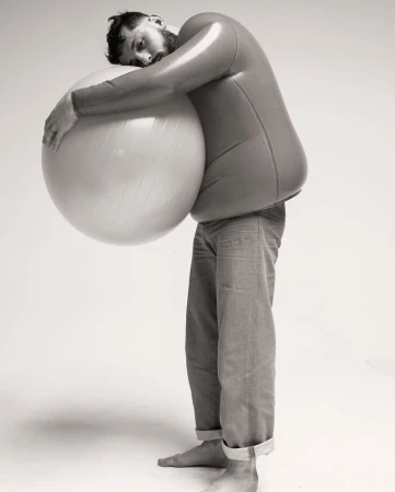 Adult male embracing exercise ball in studio setting. He is wearing a skin-tight top, jeans, and is barefoot against a plain background.