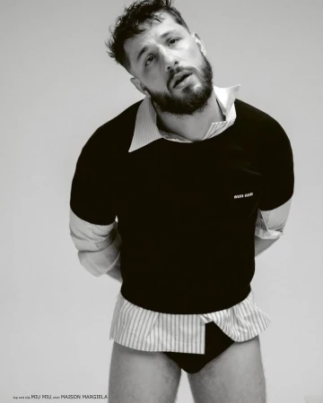 A man in black briefs and a layered black sweater and white striped collared shirt is posed in a black and white studio shot.