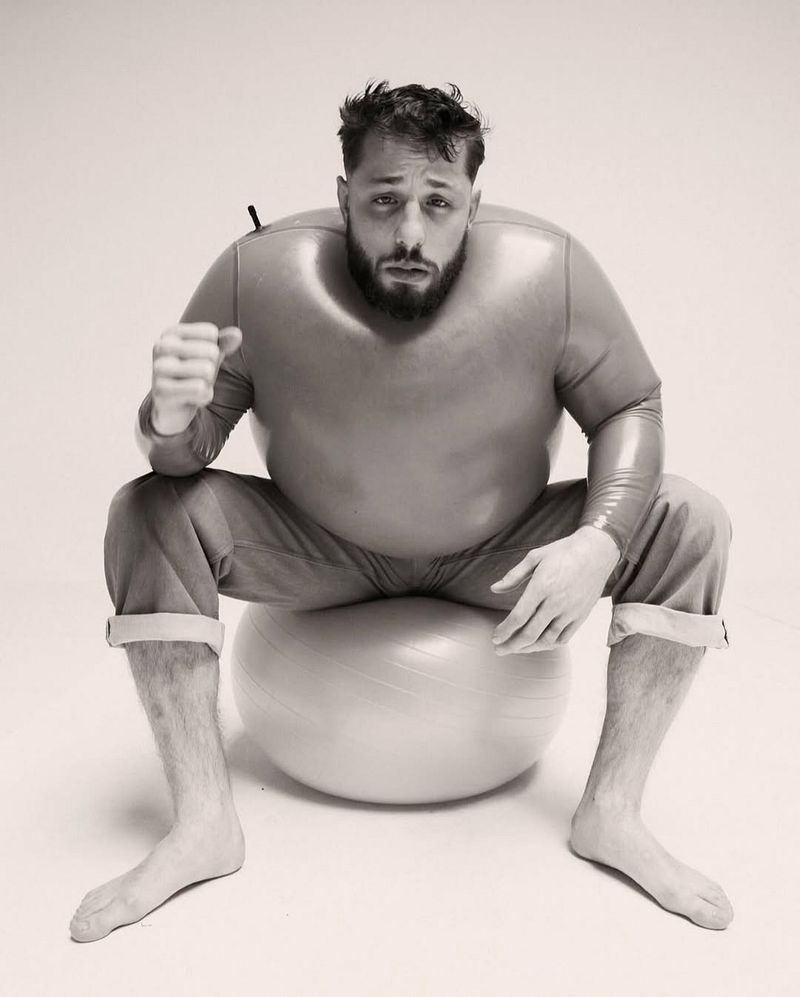 Monochrome portrait of an adult male with a beard wearing a full body suit, sitting barefoot on an exercise ball in a white studio.