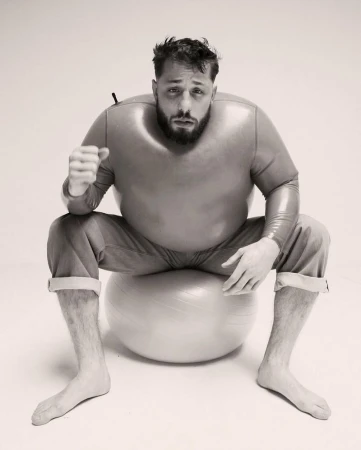 Monochrome portrait of an adult male with a beard wearing a full body suit, sitting barefoot on an exercise ball in a white studio.