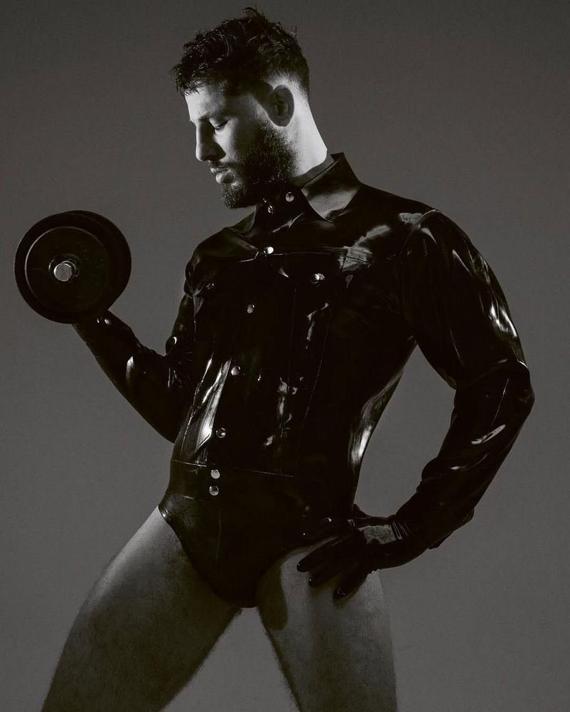 Athletic man in a shiny black bodysuit and gloves lifts a dumbbell weight in a black and white studio shot with a neutral background.